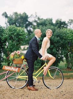 a man and woman riding on the back of a yellow bicycle with flowers in basket