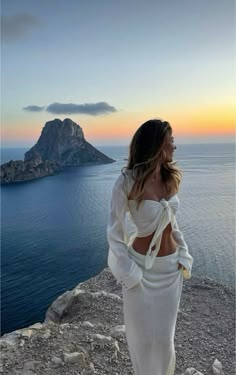 a woman standing on top of a rocky cliff next to the ocean with an island in the background