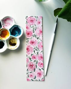 a white table topped with paint and watercolors next to a cup filled with green leaves