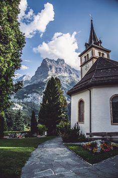 an old church with a steeple in front of a mountain range and flowers on the ground