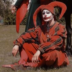 a man dressed as a clown sitting on the ground next to a black horse in a field