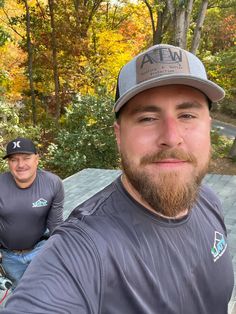 two men are taking a selfie in front of some trees and bushes, one is wearing a hat