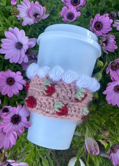 a crocheted coffee cup cozy sits in front of purple flowers and pink daisies