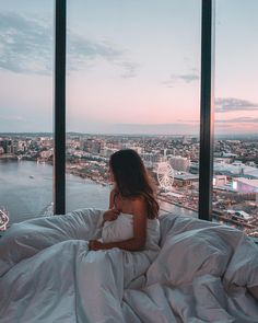 a woman sitting on top of a bed in front of a window looking out at the city