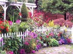 a garden filled with lots of colorful flowers next to a white picket fence and trees