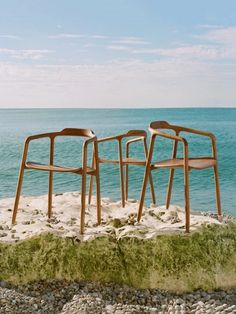 two wooden chairs sitting on top of a sandy beach next to the ocean with green grass