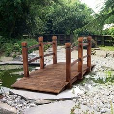 a wooden bridge over a pond surrounded by rocks and grass with trees in the background