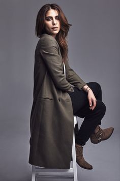 a woman sitting on top of a white chair wearing a long coat and black pants