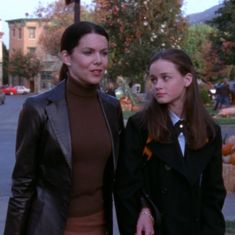 two women are walking down the street in front of some buildings and trees, one is wearing a brown turtle neck sweater