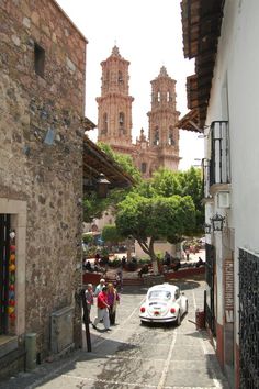 a white car driving down a narrow street next to tall buildings with towers in the background