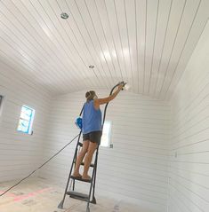 a woman is painting the ceiling in a white room with no one around her and she is on a ladder