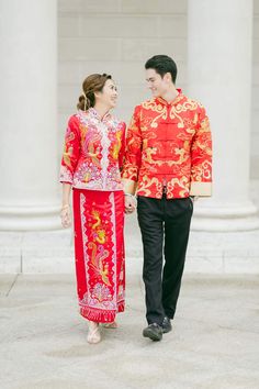 a man and woman dressed in traditional chinese clothing walking down the street with columns behind them