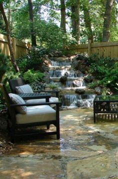 two chairs and a table in the middle of a garden with a waterfall behind them