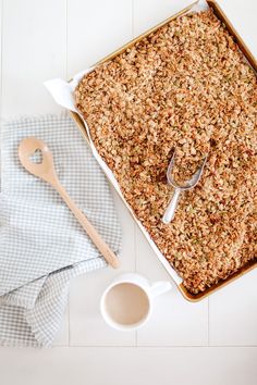 a baking pan filled with granola next to a cup of coffee and spoons