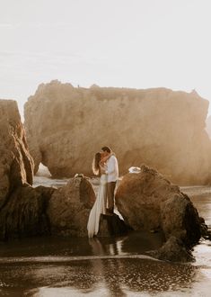 a man and woman standing on rocks in the water near some large rocks with their arms around each other