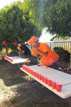 a man dressed in an orange tutu is playing with cups on a long table