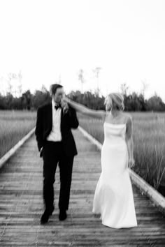 a bride and groom walking across a wooden bridge in the middle of a field with tall grass