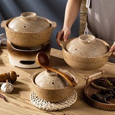 a person holding a wooden spoon over some pots and pans on a table with other items around it