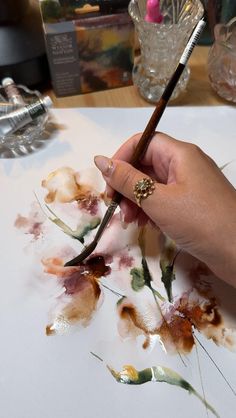 a person holding a brush in their hand while painting flowers on a white paper with watercolors