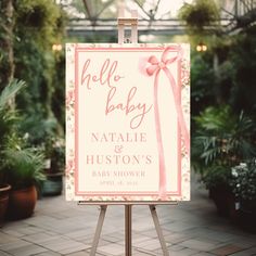 an easel with a sign that says hello baby and pink ribbon on it in front of potted plants