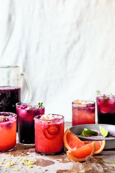 several glasses filled with different colored drinks on top of a wooden table next to sliced oranges