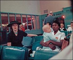 people sitting in seats at an airport with one woman holding a baby and the other man pointing