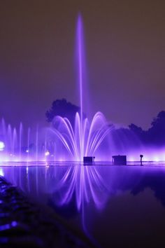 the water fountain is lit up with purple lights and reflecting in the water at night