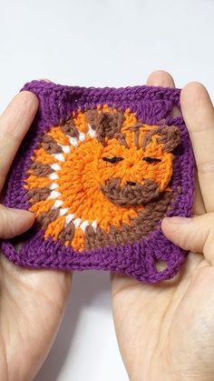 two hands holding a small crocheted square with an orange and brown face on it