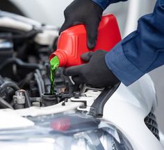 a person in black gloves is holding a red and green fuel can while pouring it into a white car's engine