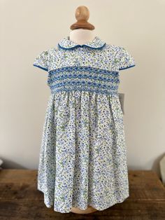 Brand: Artesania Filita Product: Nuria Composition: 100% cotton Intended Age: Toddler Description: This short sleeve, floral blue dress has a Peter Pan collar with piping (as well as on the sleeves. It is perfect for a special occasion. It fastens in the back with buttons and is fully lined. The smocked area on the chest is done in a blue pattern. Size Guide: Artisanal item- True to size. Short Sleeve Dress With Smocked Bodice For Garden Party, Garden Party Dress With Smocked Bodice And Short Sleeves, Fitted Smock Floral Dress For Spring, Fitted Short Sleeve Dress For Spring Garden Party, Floral Dress With Smocked Bodice For Garden Party, Short Sleeve Smocked Dress With Floral Print For Spring, Spring Smocked Dress With Short Sleeves And Floral Print, Spring Floral Print Smocked Dress With Short Sleeves, Short Sleeve Smocked Dress With Floral Print