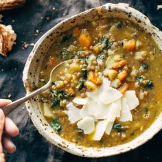 a person holding a spoon over a bowl of soup with bread on the table behind it