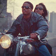 a man and woman riding on the back of a motorcycle down a street at sunset