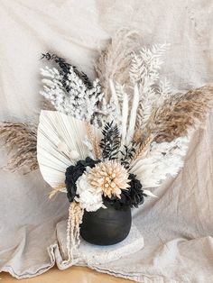 an arrangement of flowers and feathers in a black vase on a white cloth covered surface