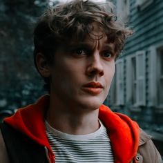 a young man with curly hair and an orange hoodie looks at the camera while standing in front of a building