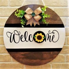 a welcome sign hanging on the side of a brick wall with a bow and sunflower