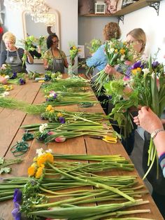 a group of women standing around a wooden table with flowers on it and people in the background