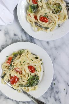 two plates of pasta with tomatoes, broccoli and spinach on a marble table