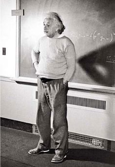 an old man standing in front of a blackboard with writing on it and wearing a sweater