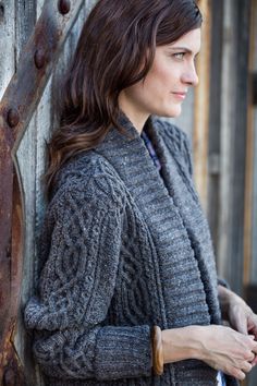 a woman leaning against a wooden wall wearing a gray cardigan sweater and blue jeans