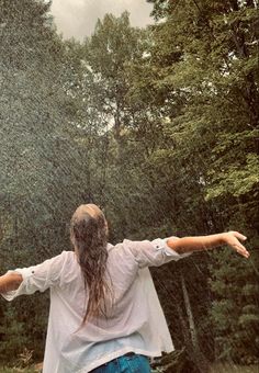 a woman standing in the rain with her arms spread out and trees behind her back