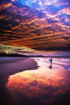 a person holding a surfboard on top of a sandy beach under a colorful sky