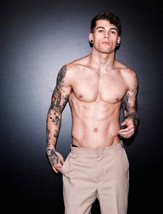 a shirtless man with tattoos standing in front of a black background and wearing khaki pants