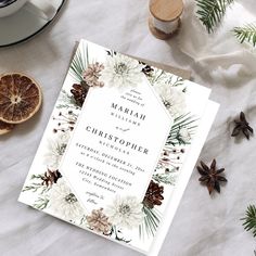 a wedding card with pine cones, oranges and evergreen leaves on the table next to it