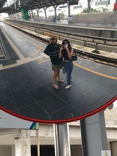 two people are standing in front of a train station with their cell phones up to their ears