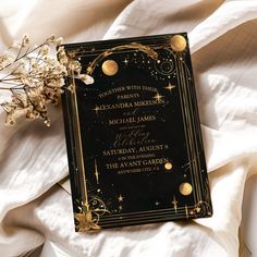 a black and gold wedding card on top of a white sheet with some dried flowers