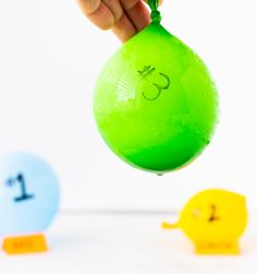 a hand is holding a green balloon in front of two small yellow ones with numbers on them