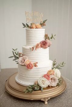 a white wedding cake with flowers and greenery on the top is sitting on a gold platter