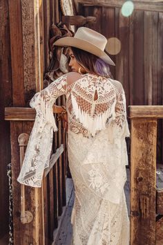 a woman in a white dress and cowboy hat leaning against a wooden fence with her back to the camera