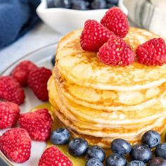 stack of pancakes with berries and syrup on white plate next to blueberries and raspberries