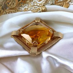 Elevate your style with this stunning vintage givre glass rhinestone and gold-plated mesh brooch, a true gem from the 1960s. The centerpiece boasts a large square-cut faceted glass stone, measuring just over 1" x 1". This mesmerizing stone showcases a captivating blend of light topaz with a deeper topaz hue at its center, all held securely in place by ornate prongs. Surrounding this remarkable stone is a square frame crafted from intricately detailed gold-plated mesh rope, featuring mesh half circles at each corner for added flair. While this piece is expertly constructed, it bears no signature, though it is reminiscent of similar creations by Hobe, known for their gold mesh designs. Resting in very good vintage condition, this brooch promises to stand the test of time. With its unique and Elegant Yellow Brooches For Party, Elegant Yellow Party Brooch, Elegant Yellow Party Brooches, Luxury Yellow Gold Brooches For Parties, Gold Jeweled Brooches For Formal Occasions, Gold Brooches With 17 Jewels For Evening, Elegant Gold Jeweled Brooches, Formal Gold Jeweled Brooches, Gold Jeweled Brooches For Party